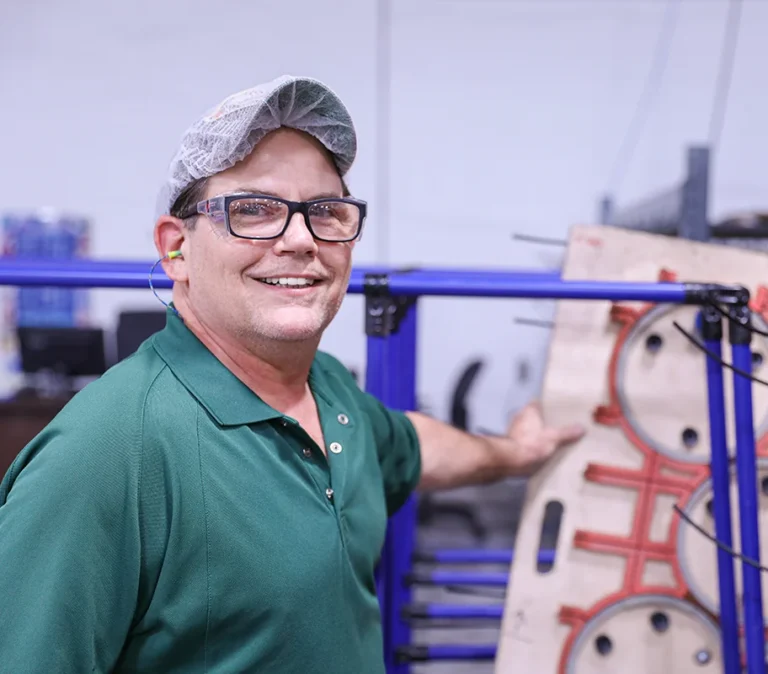 A smiling manufacturing employee wearing safety glasses, a hair net, and ear protection stands beside an equipment rack on the production floor.