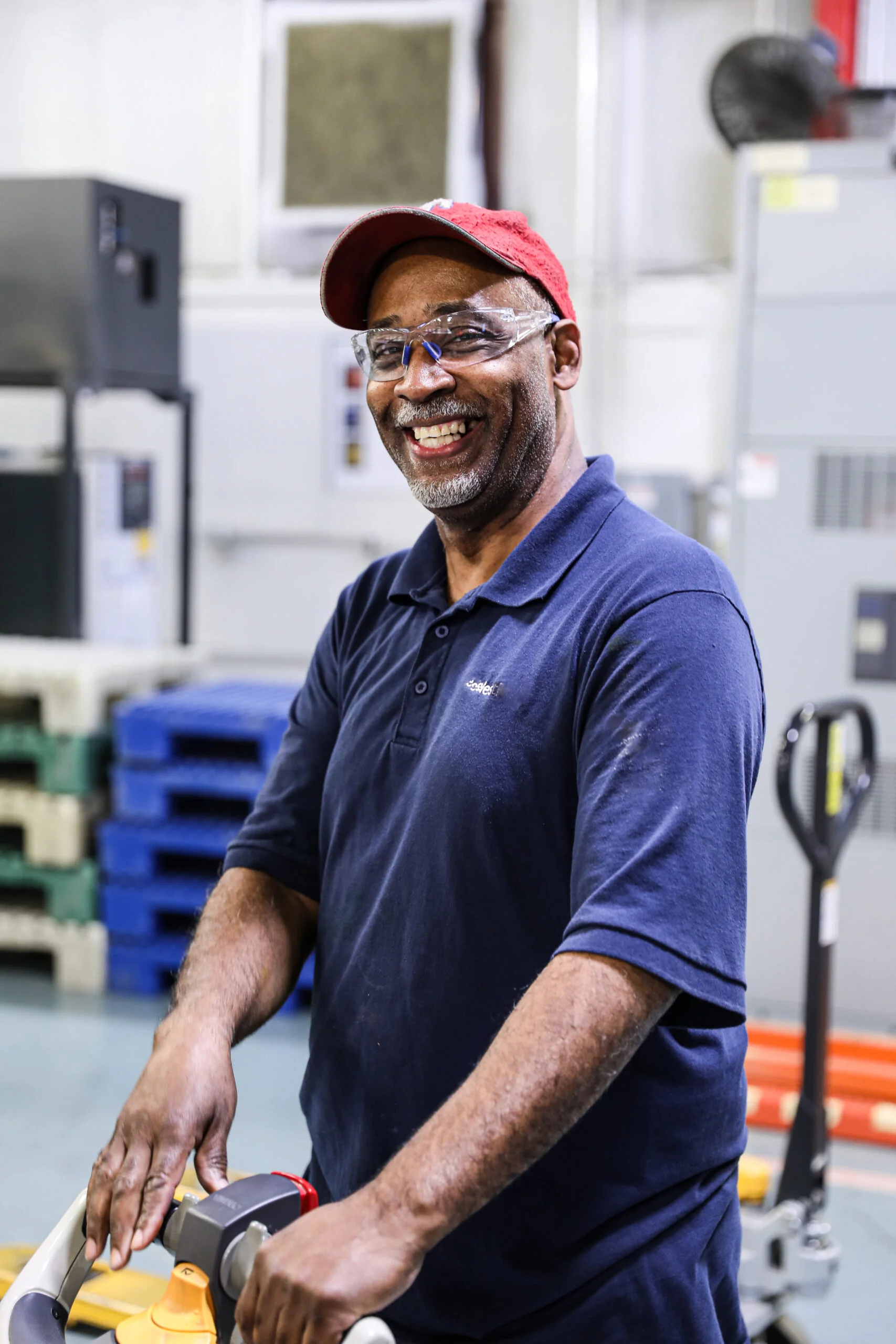 A smiling warehouse worker wearing safety glasses operates a pallet jack on the warehouse floor.
