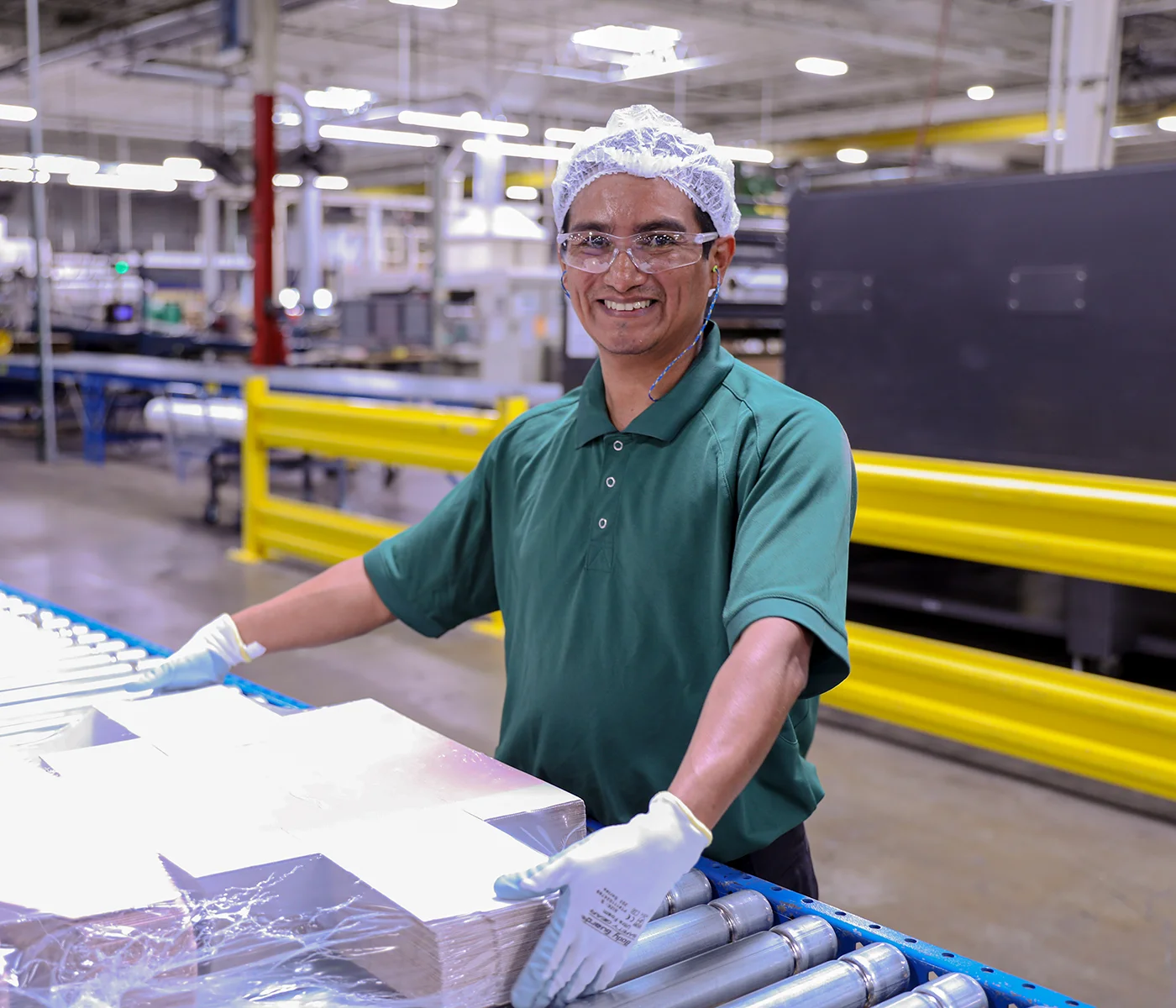 A smiling manufacturing employee wearing safety glasses, a hair net, and gloves handles materials on a conveyor line in a large production facility.